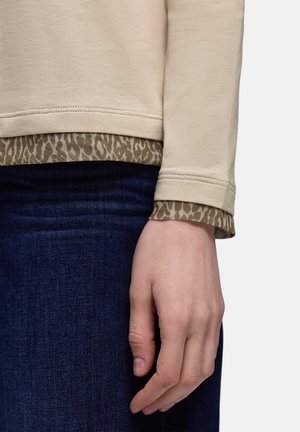 Beige långärmad topp med en strukturerad djurprintad nederkant, kombinerad med mörkblå jeans; synlig hand som vilar på denimtyget.