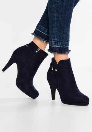 Botas de tobillo de ante azul marino con tacón alto, que cuentan con un cierre lateral y detalles en plata. Acabado texturizado y diseño de punta redonda.
