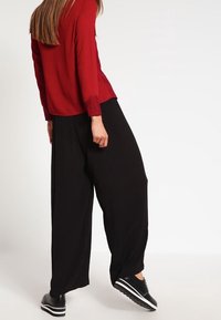 Blouse rouge à manches longues avec des accents en dentelle ; associée à un pantalon noir ample et des baskets noires avec des rayures blanches. Textures lisses partout.