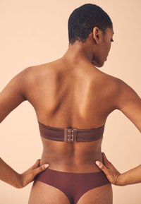 Soutien-gorge marron sans bretelles avec une fermeture à crochets au dos. Tissu lisse avec un design sans couture, porté avec des culottes marron assorties.