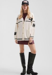 Młoda kobieta w beżowym kardiganie z łatami, bordowej sukience, czarnych wysokich kozaczkach oraz czapce z logo.