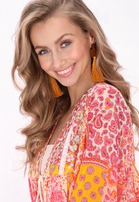 Virágmintás rózsaszín és narancssárga árnyalatú kimono, paisley mintával, csipkés szegéllyel és laza szabással. Hosszú, göndör hajjal és bojt fülbevalókkal rendelkezik.