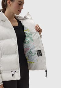 Giubbotto piumino bianco con un design trapuntato. Fodera interna con motivi astratti verdi e gialli. Top a coste nero sotto.
