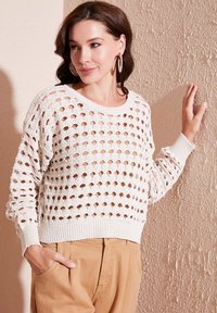 Suéter blanco de punto con grandes agujeros abiertos, mangas largas y cuello redondo. Combinado con pantalones beige y situado frente a una pared texturizada.