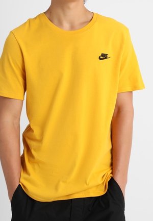Personne portant un t-shirt Nike jaune uni à manches courtes avec un logo noir sur la poitrine, les mains partiellement dans les poches d'un pantalon noir.