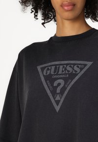 Črna oversize majica iz mehkega bombaža, s trigonalnim sivim logotipom na sredini in napisom "GUESS® ORIGINALS WASHED JEANS."