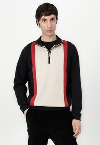 Schwarzer Pullover mit Reißverschluss am Hals, einem cremefarbenen gemusterten Mittelteil und roten seitlichen Streifen, mit gerippten Bündchen und Saum, getragen mit schwarzen Hosen.