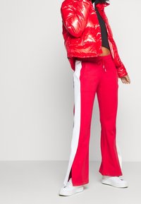 Persona che indossa un giaccone ammortizzato rosso lucido, pantaloni da track rossi con strisce bianche sui lati e sneakers bianche, in piedi su uno sfondo chiaro e semplice.
