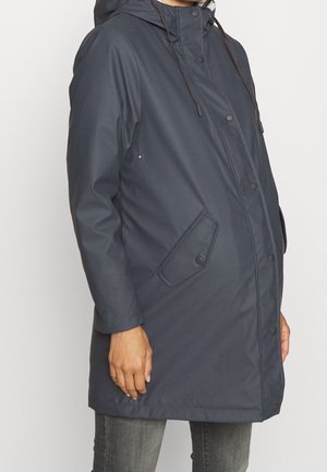 Personne portant un imperméable à capuche bleu foncé avec boutons-pression et poches avant à rabat, debout devant un fond clair et uni.