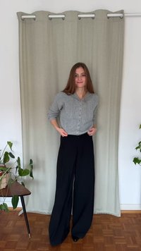 Szary kardigan na guziki o prążkowanej fakturze, zestawiony z szerokimi czarnymi spodniami. W tle znajduje się jednolita zasłona i roślina doniczkowa.