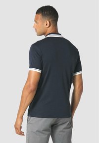 Polo verde navy con colletto a righe bianche e orli delle maniche bianchi. La vista posteriore mostra maniche corte e una texture liscia in cotone.