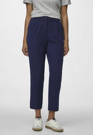 Pantaloni tapered blu navy con pieghe frontali, chiusura a bottone e tasche laterali, abbinati a sneaker bianche con dettagli grigi.
