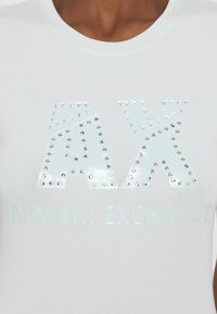 Balta medvilninė marškinėlių su blizgiu iškilmingu "AX" logotipu, papuoštu kristalais, ir užrašu "ARMANI EXCHANGE" apačioje.