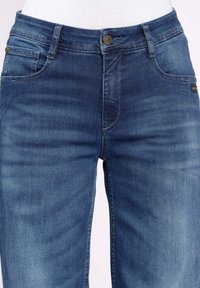 Jeans de mezclilla en azul oscuro con una textura lisa, cinco bolsillos, cremallera en la bragueta y un botón de latón en la cintura. Áreas sutilmente desgastadas en la tela.