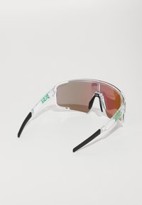 Przezroczyste sportowe okulary przeciwsłoneczne z brązowymi soczewkami, czarnymi gumowymi końcówkami na uszy i zielonymi logo na zausznikach. Smukły design, owinięty kształt.