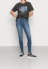 Camiseta negra con un estampado de Van Gogh, jeans ajustados azules y zapatillas de skate negras con suelas blancas, de pie contra un fondo neutro.