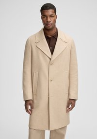 Cappotto in lana beige con collo a risvolto, cinque bottoni, due tasche laterali e vestibilità morbida, indossato sopra una maglietta polo marrone scuro.