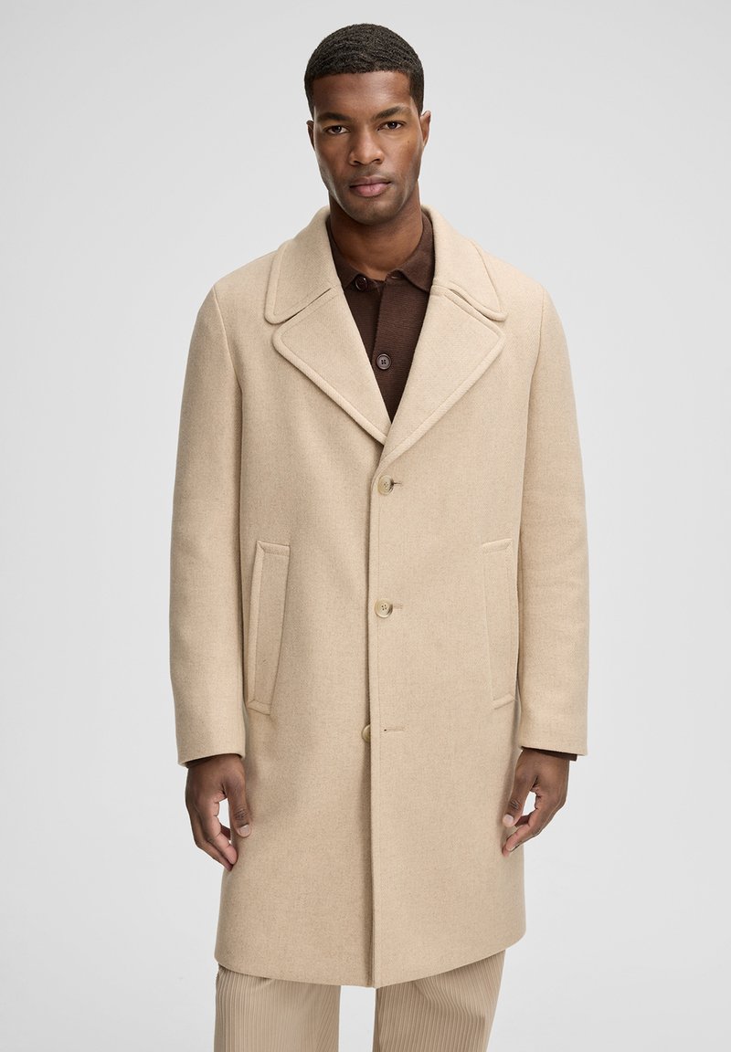 Cappotto in lana beige con collo a risvolto, cinque bottoni, due tasche laterali e vestibilità morbida, indossato sopra una maglietta polo marrone scuro.