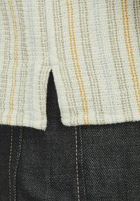 Vævet top i et struktureret, lysekræmmet stof med striber i lyseblå, grå og gul. Sømmen har et lille slidser; båret over mørke jeans.