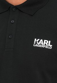 Črna polo majica s tremi gumbi in belim logotipom "KARL LAGERFELD" na prsih, ki vsebuje majhen grafični obris črke K.