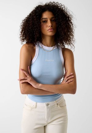 Vrouw met krullend haar, gekleed in een lichtblauw mouwloos topje en witte spijkerbroek, staand met gekruiste armen tegen een effen achtergrond.