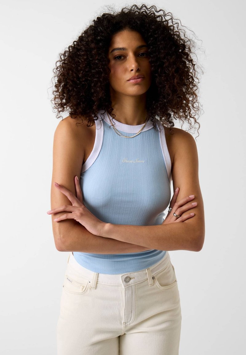 Vrouw met krullend haar, gekleed in een lichtblauw mouwloos topje en witte spijkerbroek, staand met gekruiste armen tegen een effen achtergrond.