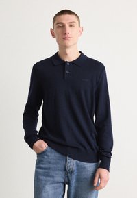 Sötétkék, hosszú ujjú pólóing klasszikus gallérral, gombos nyitással és bordázott mandzsettával. Diszkrét logó a mellrészen. Világos farmernadrággal viselve.