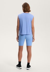 Světle modrý top bez rukávů s volným střihem, v kombinaci se světle modrými šortkami. Oblečení má hladkou texturu a minimalistický design. Černá obuv s bílou podrážkou.