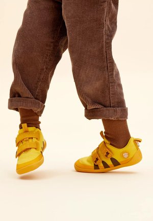 Kind trägt braune Cordhosen und gelbe Sneaker mit Klettverschlüssen, steht auf einem hellbeigen Boden.