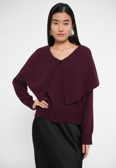 Maglione lavorato a maglia bordeaux con un design ampio e drappeggiato e scollo a V, caratterizzato da polsini e orlo a coste. Abbinato a una gonna nera.