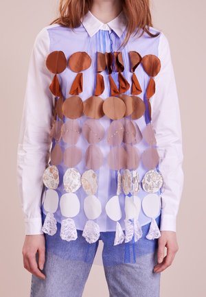 Osoba ubrana w białą, zapinaną na guziki koszulę pod przezroczystą niebieską kamizelką ozdobioną rzędami dużych, brązowych, beżowych i białych okrągłych aplikacji z tkaniny.