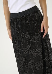 Černá maxi sukně s flitry, která má texturovaný, třpytivý povrch. Má široký elastický pas a vrstvený design pro komfortní pohyb a splývání.