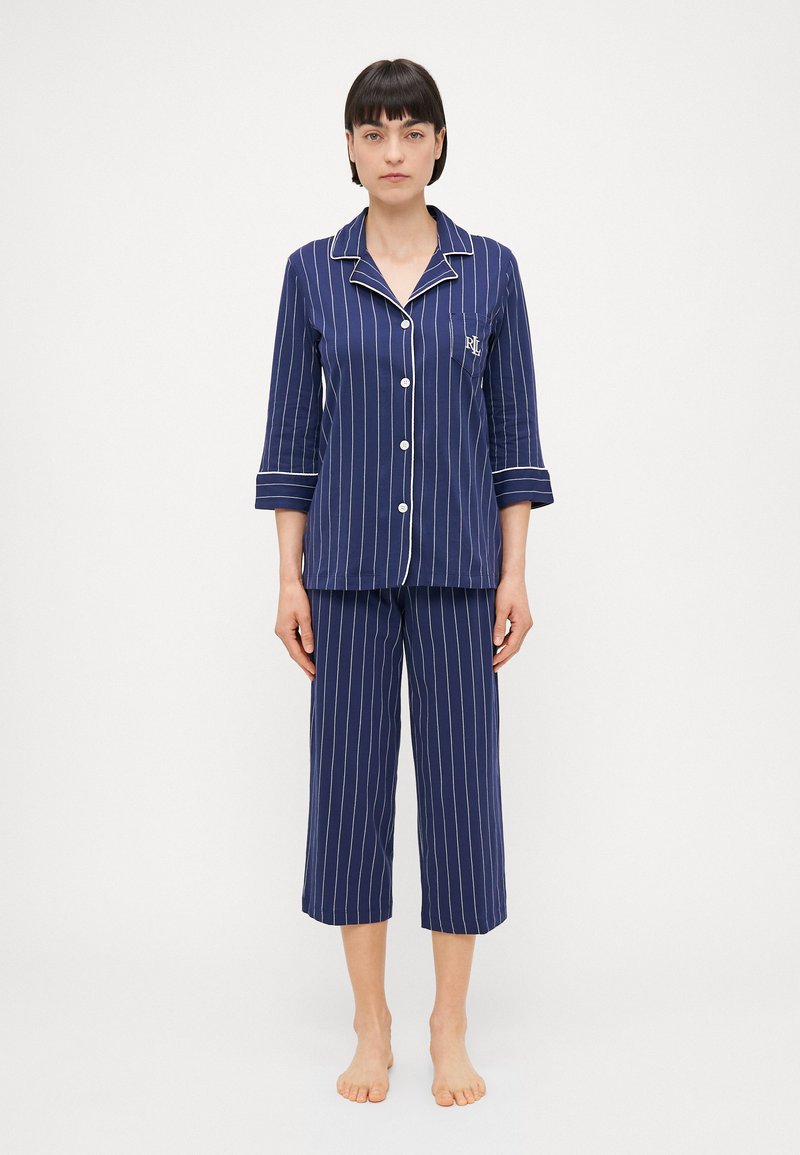 Conjunto de pijama listrado em azul-marinho, com uma camisa de botões de mangas três-quartos e calças cropped combinando; inclui detalhes brancos e um bolso.