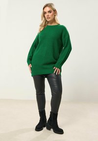 Zielony sweter dzianinowy z szerokimi rękawami i luźnym krojem, w połączeniu z czarnymi skórzanymi legginsami i czarnymi botkami na obcasie. Gładka tekstura w całej powierzchni.