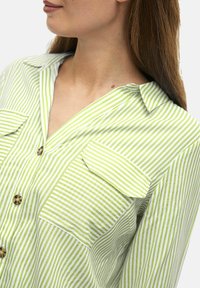 Camisa de rayas verdes y blancas con collar, dos bolsillos frontales y cierre de botones. Confeccionada con una tela ligera de textura suave.