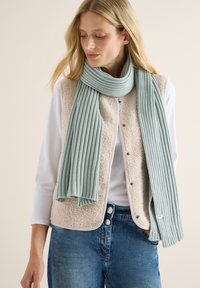 Beige fleece vest med trykknapper, parret med et ribbet mintgrønt tørklæde. Modellen har en hvid langærmet top og blå jeans på.