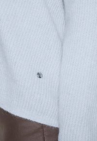Pull blanc doux en tricot avec un petit logo métallique rond près de l’ourlet, associé à un pantalon marron.