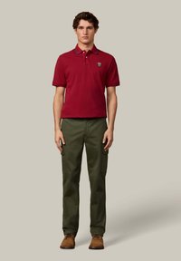 Bordeauxrode polo met kraag en embleem, gecombineerd met olijfgroene cargobroek. Model draagt bruine schoenen. Eenvoudig, aangesloten ontwerp.