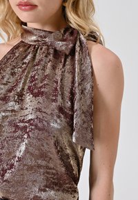 Top halter in tessuto metallico color bordeaux e oro, con un grande fiocco al collo e una superficie morbida e testurizzata con una finitura scintillante.