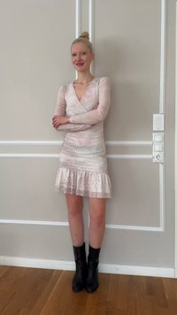 Ung kvinne med blondt hår i en knute, iført en lyserosa kjole med mønster og svarte ankelstøvler, står med armene i kors innendørs.