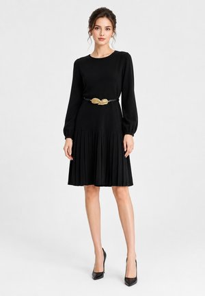 Femme portant une robe noire à manches longues avec une jupe plissée, une ceinture feuille d'or et des talons hauts noirs, debout devant un fond uni.