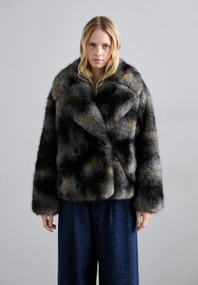 Kobieta z długimi blond włosami, ubrana w wzorzystą, ciemnoszaro-czarną kurtkę z sztucznego futra i luźne ciemnoniebieskie spodnie na jednolitym tle.