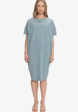 Kobieta nosząca luźną, sięgającą do kolan, jasnoniebieską sukienkę z krótkimi rękawami, beżowe sandały na odkryte palce oraz złoty naszyjnik z wisiorkiem, zwrócona twarzą do przodu.