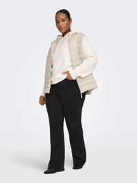 Beige vadderad väst över en vit huvtröja, kombinerad med svarta utsvängda jeans. Västen har en quiltad design och framfickor. Svarta skor fullbordar looken.