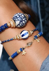 Trois bracelets en perles avec des motifs bleu et blanc, des accents dorés et diverses formes, y compris des designs ronds et floraux. Des perles texturées sont incluses.