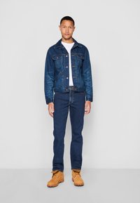 Hombre de pie contra un fondo liso, vistiendo una chaqueta de mezclilla azul, jeans a juego, camiseta blanca y botas de trabajo marrones.