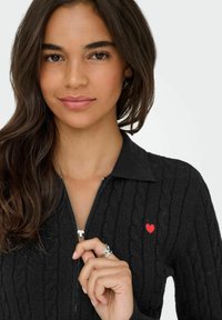 Czarny sweter zapinany na zamek, z plecionym wzorem, z małym haftem czerwonego serca po lewej stronie klatki piersiowej.