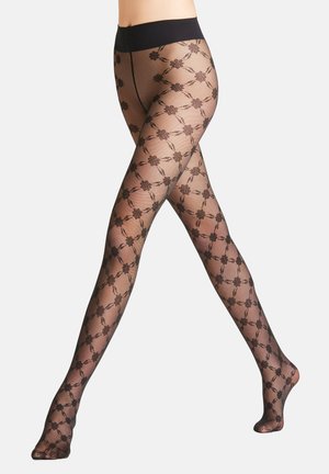 Legs crossed wearing sheer black patterned tights with floral design and solid black waistband on a plain white background.