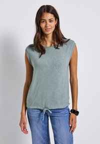 Jasnozielony, bezrękawnik z okrągłym dekoltem, miękka tekstura i wiązanie u dołu, połączony z niebieskimi dżinsami.