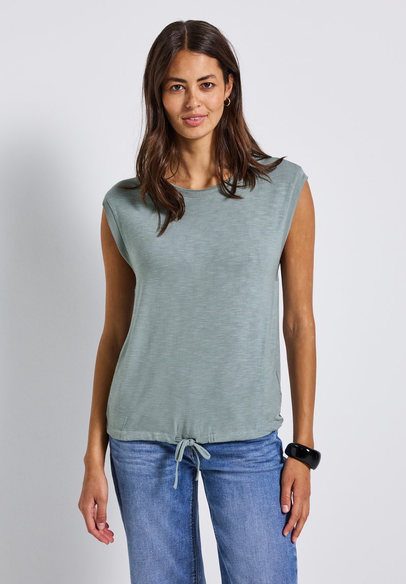 Jasnozielony, bezrękawnik z okrągłym dekoltem, miękka tekstura i wiązanie u dołu, połączony z niebieskimi dżinsami.